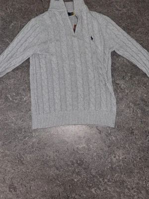 Ljusgrå kabelstickad tröja Polo Ralph Lauren - Ljusgrå kabelstickad tröja från Polo Ralph Lauren med hög krage och dragkedja. Tröjan har det klassiska broderade logotypen på bröstet och ribbade muddar vid ärmslut och nederkant. Perfekt för dig som gillar en stilren och tidlös look.