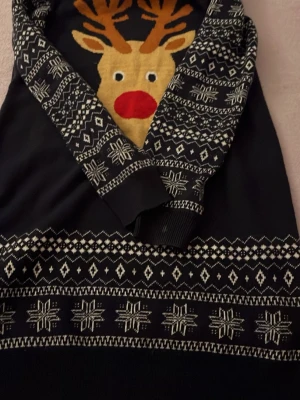 Svart jultröja med renmotiv - Mysig svart stickad tröja med stort renmotiv framtill och vita snöflingor samt geometriska mönster på ärmar och nederkant. Perfekt för julens alla tillfällen och ger en härlig vinterkänsla. Tröjan har långa ärmar och ribbade muddar.