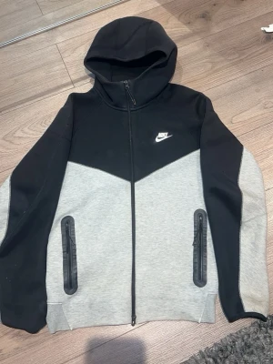 Nike hoodie svart och grå M - Nike hoodie i svart och grått med dragkedja och två fickor med svarta detaljer. Klassisk Nike-logga på bröstet och huva. Perfekt för en sportig och avslappnad stil.
