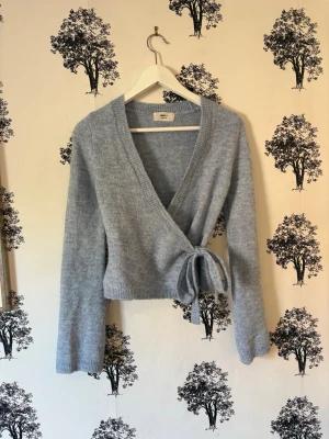 Ljusblå omlottkofta i mjuk stickning - Supersnygg ljusblå omlottkofta i mjuk stickad kvalitet. Koftan har långa ärmar, bred ribbad mudd och knyts i sidan för en schysst passform. Perfekt att slänga över en topp för en chill men ändå stilsäker look.