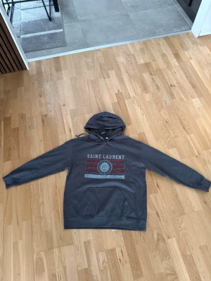 Mörkgrå Saint Laurent hoodie - Snygg mörkgrå hoodie från Saint Laurent med tryck framtill i vitt, rött och blått. Klassisk huva med dragsko och stor magficka. Perfekt för dig som vill ha en stilren och trendig look med streetwear-vibe. Är Xl men sitter mer som L. Ni vet varför den är billig🤝