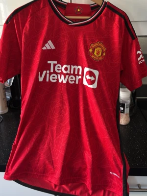 Manchester United matchtröja röd - Säljer en röd Manchester United matchtröja från Adidas med klubbmärke och TeamViewer-logga på bröstet. Tröjan har svarta detaljer på axlar och sidor, samt vita Adidas-loggan. Tillverkad i lätt och ventilerande material, perfekt för fotboll eller träning.