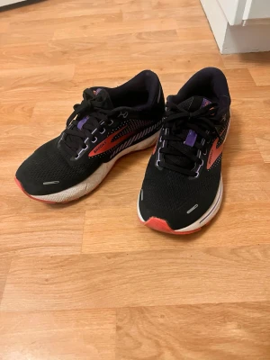 Svarta Brooks sneakers med lila detaljer - Snygga svarta sneakers från Brooks med lila och korallfärgade detaljer. Skorna har en låg siluett, svart mesh-överdel och vita sulor med inslag av korall. Perfekta för dig som tycker om att springa eller träna!! Nypris runt 1700kr
