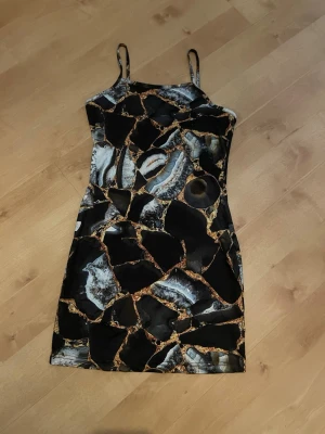 Svart mönstrad kort klänning - Svart kort klänning med unikt mönster i grått, vitt och guld. Klänningen har smala axelband och figurnära passform. 