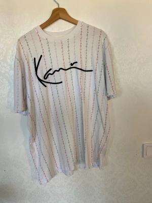 Vit t-shirt från Karl Kani med tryck - Säljer en vit t-shirt från Karl Kani med färgglada vertikala ränder och stort svart logotyptryck på bröstet. T-shirten har en relaxed fit och är gjord i mjuk bomull. Perfekt för dig som gillar streetwear och vill sticka ut lite extra.
