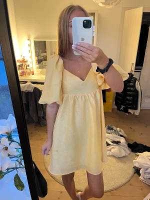 Gul kort klänning med puffärm - Söt gul kort klänning med v-ringning och stora puffärmar. Klänningen har en lätt struktur i tyget och en flowy passform som ger en romantisk vibe. Perfekt för varma dagar och somriga tillfällen.