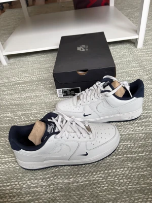 Nike Air Force 1 vita/navy sneakers -  NYA. Säljer ett par fräscha Nike Air Force 1 sneakers i vitt läder med mörkblå detaljer och innersula. Klassisk siluett med perforerad tå, vita skosnören och diskret mörkblå mini-swoosh på sidan. Perfekta för dig som gillar stilrena och tidlösa sneakers.