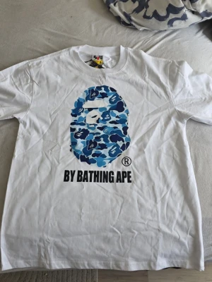 Vit BAPE t-shirt med blått camotryck - Säljer en vit t-shirt från BAPE med det klassiska apansiktet i blått camomönster på bröstet och texten 'BY BATHING APE' under. T-shirten har rund hals och korta ärmar. Perfekt för dig som gillar streetwear och vill sticka ut.