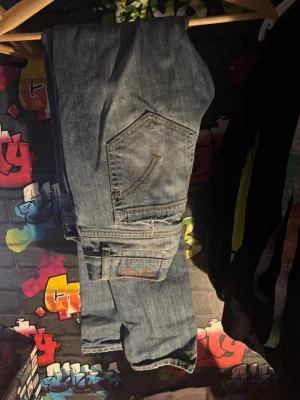 Blå slitna jeans med fickdetaljer - Säljer ett par blå jeans med slitningar och coola fickdetaljer. Jeansen har en avslappnad passform och dekorativa sömmar på bakfickorna. Perfekta för en casual streetwear-look och tillverkade i klassisk denim.