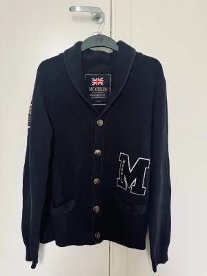 Marinblå kofta från Morris - Snygg marinblå kofta från Morris med stora knappar och två fickor framtill. Koftan har en stor vit M-patch på ena sidan och en krage som ger en klassisk collegestil. 