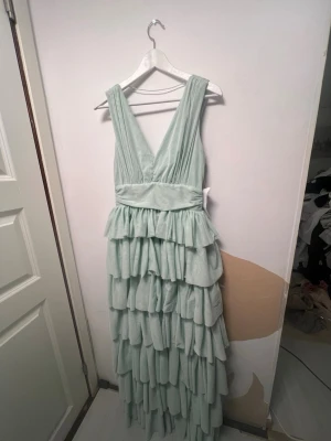Ljusgrön långklänning med volanger - Säljer en ljusgrön långklänning med flera lager volanger och djup v-ringning. Klänningen har breda axelband och markerad midja, vilket ger en elegant siluett. Tillverkad i tunn, luftig polyester som ger ett drömmigt intryck. Balklänning 