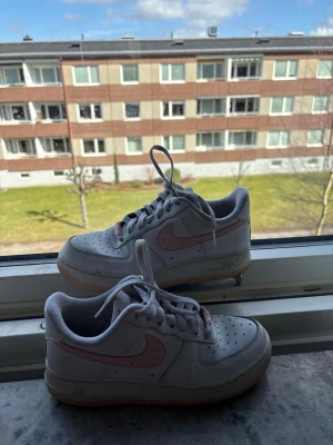 Nike Air Force 1  - Snygga Nike Air Force 1 sneakers i vitt läder med rosa swoosh och detaljer. Klassisk siluett med perforerad tå och gummisula i ljusbrun färg. Perfekta för dig som vill ha en stilren och trendig look med en touch av färg.