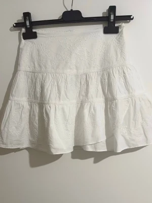 Vit broderad volangkjol från Zara  - Söt vit kjol med broderade blommor och volanger. Kjolen har resår i midjan baktill för extra komfort och är gjord i ett lätt bomullstyg. Perfekt för en somrig och fräsch look.