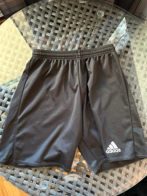 Svarta träningsshorts från Adidas - Säljer ett par svarta träningsshorts från Adidas med klassisk logga broderad på vänster ben. Shortsen har elastisk midja och är tillverkade i ett lätt och svalt polyestermaterial, perfekta för sport och aktivitet.