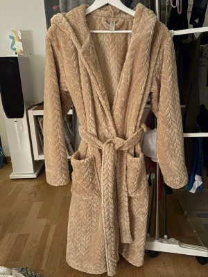 Beige fluffig morgonrock med huva - Supermjuk beige morgonrock med huva. Perfekt för att mysa runt hemma eller bara att ha på morgonen Tillverkad i fluffigt, värmande material som känns riktigt skönt mot huden.