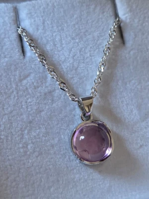 Silverhalsband med lila sten - Snyggt halsband i silverfärgad metall med tvinnad kedja och ett runt hänge med en glansig, lila sten. Hänget har en mjuk, rundad form och ger ett elegant intryck. Perfekt accessoar för att lyfta din outfit med en färgklick.