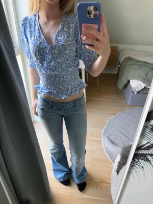 Blommig blå volangblus - Söt blus i ljusblått med vitt blommönster och volangdetaljer. Modellen har v-ringning, smockad midja och trekvartsärm med puff. Tunn och luftig, perfekt till jeans eller kjol. Materialet känns som viskos eller polyester.