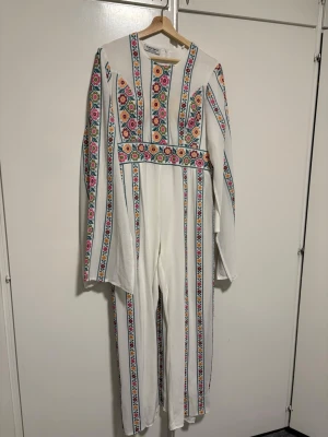 Palestinsk Klänning  - En helt ny unik vit jumpsuit med långa ärmar och färgglada blommiga broderier i ränder längs hela plagget. Rund halsringning och markerad midja med broderat bälte. Perfekt för dig som vill sticka ut med en bohemisk och retroinspirerad stil. 