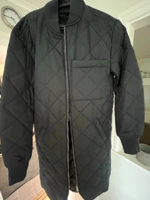 Svart lång pufferjacka från 157 - Snygg svart lång pufferjacka från 157 med quiltat mönster och rak passform. Jackan har en stor bröstficka, ribbade muddar och dragkedja framtill. Perfekt för kalla dagar och ger en clean look.