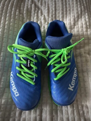 Blåa Kempa inomhusskor med gröna detaljer - Säljer ett par sportiga blåa inomhusskor från Kempa med gröna snören och detaljer. Skorna har en låg profil, ventilerande meshmaterial och slitstark sula som passar perfekt för handboll eller andra inomhussporter. Snygg design med Kempa-logga på sidan. Kan mötas upp i Sthlm