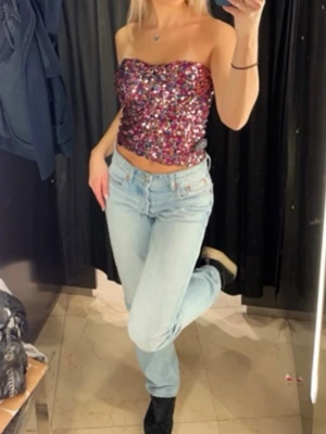 tubtopp glittrig  - Säljer en glittrig tubtopp täckt av paljetter i rosa, lila och guldtoner. Toppen är croppad och axelbandslös. Sitter tight och har en snygg bandeauformad passning. Supersnygg till både jeans och kjol!