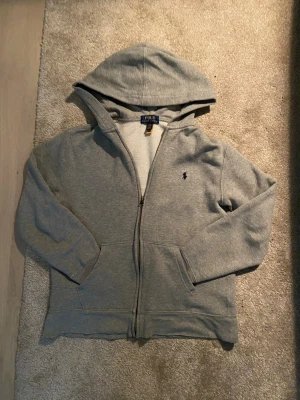 Ralph Lauren Zip Hoodie - Grå zip-hoodie från Ralph Lauren 🐎 Storlek L (14–16)  • Supermjuk insida • Klassisk liten logga • Lite småskador som man ser på bilderna - annars bra skick.  Säljer då den inte används längre. Passar till allt – fett clean basic 👌 