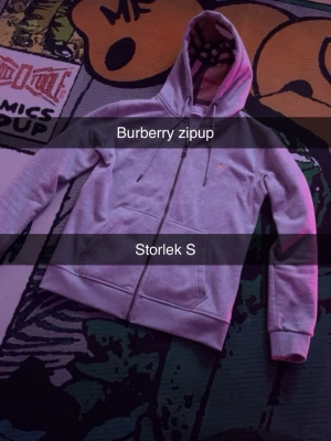 Grå zip hoodie från Burberry - Säljer en grå zip hoodie från Burberry med klassiskt rutigt foder i huvan och två fickor framtill. Dragkedja hela vägen och diskret Burberry-logga på bröstet. Perfekt för dig som gillar stilrena och exklusiva plagg.