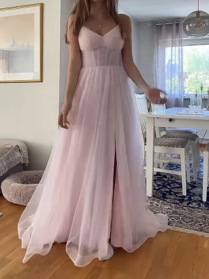 Ljusrosa långklänning med slits Balklänning - Romantisk ljusrosa balklänning med tunna axelband och v-ringad urringning. Klänningen har ett lager av tunn tyll över ett mjukt foder och en elegant slits framtill. Perfekt för bal. Sitter figurnära upptill och faller mjukt från midjan. Storlek 34, JJ’ house, nypris 1500. Aldrig använd