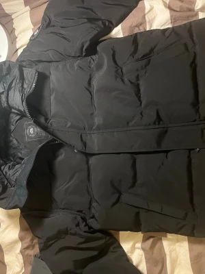 Svart pufferjacka från Canada Goose - Säljer en svart pufferjacka från Canada Goose i storlek M. Jackan har en stilren look med broderad logga på ärmen, dragkedja framtill och två fickor. Materialet är polyester och dun, perfekt för kalla dagar. Jackan har en normal passform och långärmade ärmar.