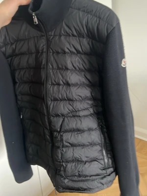 Svart cardigan från Moncler - (Förlåt för otydliga bilder). Svart cardigan från Moncler med quiltad framsida i glansigt material och stickade ärmar samt rygg. Jackan har hög krage, två dragkedjefickor och Moncler-logga på ärmen. Perfekt för kalla vårdagar och snygg att matcha med jeans.