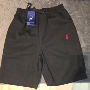 Svarta shorts från Polo Ralph Lauren - Snygga svarta shorts från Polo Ralph Lauren med elastisk midja och snörning. Klassisk logga broderad i vitt på ena benet. Perfekta för en avslappnad stil och tillverkade i mjuk bomull för extra komfort.