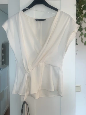 Vit omlottopp från Zara - Snygg vit omlottopp från Zara med v-ringning och lätt volang nertill. Toppen är ärmlös och har en luftig, feminin passform som passar perfekt till jeans eller kjol. Toppen har tyvärr inte kommit till användning, därför den säljs