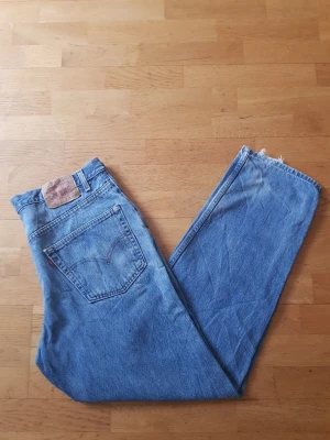 Levi's 550 blå raka jeans - Klassiska blå Levi's 550 jeans med rak passform och hög midja. Jeansen har fem fickor, bälteshällor och den ikoniska läderlappen bak i linningen. Tillverkade i slitstark denim med vintagekänsla och rå kant vid benslut. Perfekta för en avslappnad och tidlös stil.