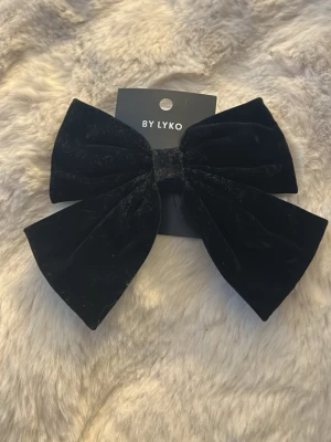 Svart sammetsrosett från Lyko - Stilren svart hårrosett i mjuk sammet från Lyko. Rosetten har en klassisk form och sitter på ett metallspänne som gör den enkel att fästa i håret. Perfekt accessoar för att lyfta din look med en elegant touch.