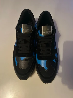 Valentino blå camo sneakers - Snygga sneakers från Valentino med blå och svart camo-mönster. Skorna har svarta snören, svart sula och en klassisk låg siluett. Märkeslogga på plösen och cool design som sticker ut. Perfekta för dig som gillar streetwear och vill ha något unikt.
