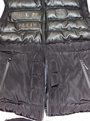Svart dunväst från Philipp Plein - Snygg svart dunväst från Philipp Plein med glansiga partier upptill och tydlig logga på bröstet. Västen har två dragkedjeförsedda fickor och coola detaljer i metall. Perfekt för lager på lager och streetwear-stil. Har haft den i 1 år men den är som ny 
