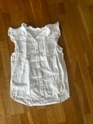 Vit ärmlös blus med volangkant - Söt vit blus i luftig bomull med knappar framtill och volangkant vid ärmhålen. Blusen har en lätt rynkad detalj vid axlarna och en mjukt rundad nederkant. Perfekt för en avslappnad och somrig look.