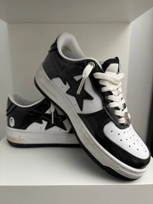 Svarta och vita BAPE STA sneakers - Säljer ett par BAPE STA sneakers i svart och vitt med glansig finish. Skorna har den ikoniska stjärnloggan på sidan, vita snören med metalltips och perforerad tåbox. Yttermaterialet är lackat läder och sulan är vit med svart undersida. Perfekt för dig som gillar streetwear.
