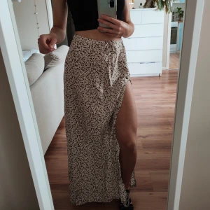 Långkjol med slits  - Långkjol med slits. Med vita, beiga och svarta blommor. Står att det är storlek L men tack vare knytning så kan man justera. Finns även en dragkedja för enklare på och avtagning.