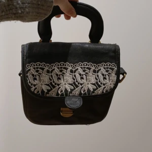 En svart liten armväska  - En liten svart armväska, lite oklart vart i från. Är lite svårt att stänge lite trastig men har ett jätte fint vitt mönster.