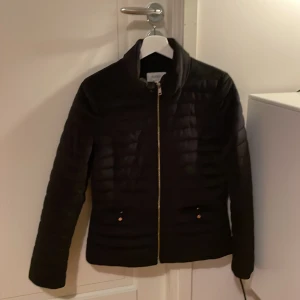 Höstjacka i mjukt material  - Svart kortare jacka, lätt fodrad i mjuk polyester. Lite sammetskänsla på yttertyget samt guldkedja och guldknappar +finns extra knappar. Lite använd och normal till storleken. Väldigt fint skick. Nypris 1000kr.