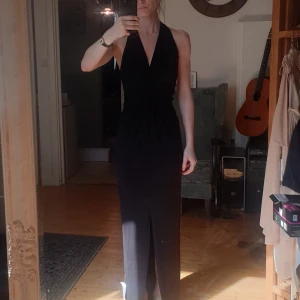 Långklänning - Svart maxiklänning med halterneck och tvinnad framtill. Öppen rygg. Slits.  Ny och aldrig använd. Lappar kvar.