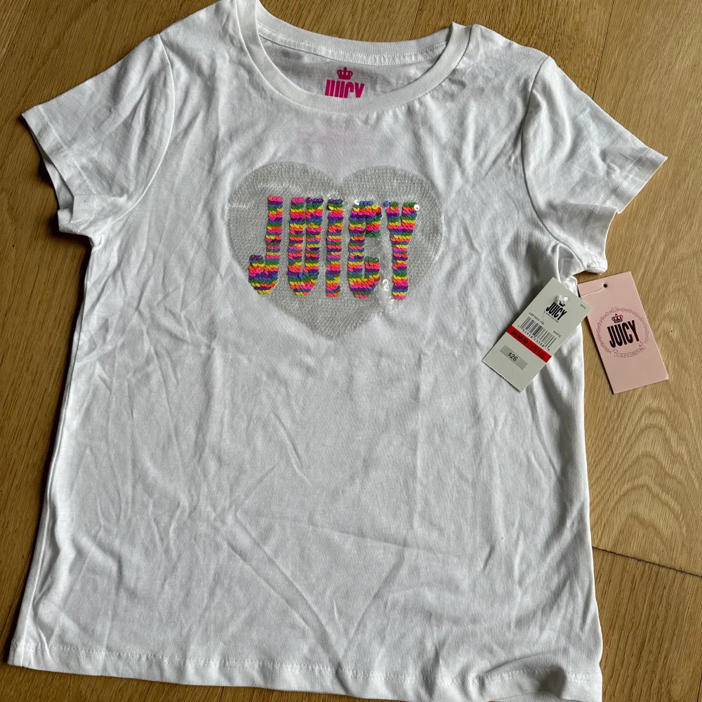 Vit Juicy t-shirt - aldrig använd med prislappen på. Vändbar paljett text ’Juicy’ som ändrar färg. Köpt i USA ($26). Vi köpte flera färger och av någon anledning blev den vita aldrig använd. . T-shirts.