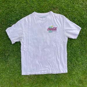 En t-shirt i en vit färg med svag nyans av rosa i. Coca cola tryck på bröstet samt ryggen, made in portugal. 