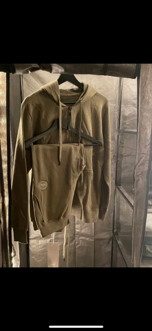 Goose dress - Splitter ny Grön tracksuit från Canada Goose. Tillverkad av 100% bomull. Justerbar luva med dragsko, ribbstickad fåll och ärmslut och två handfickor framtill. Rund patch med logtoyp på vänster ärm. Samt Patch på vänsta sidan  På byxan 