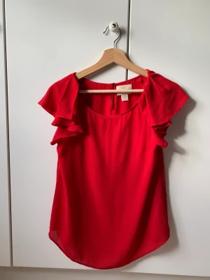 Röd Blus - Röd blus med volanger på axlarna och en liten knapp i nacken. Materialet är inte genomskinligt men tunnt och luftigt!