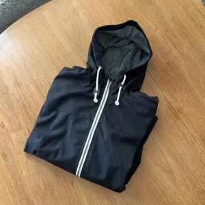 Stilren vindjacka (svart) - En i princip oanvänd jacka från Brave Souls. Stilren i svart färg med vita detaljer. Samt bruna detaljer på snören. Kragen är något högre för att ge bättre skydd mot vind. Storlek medium.