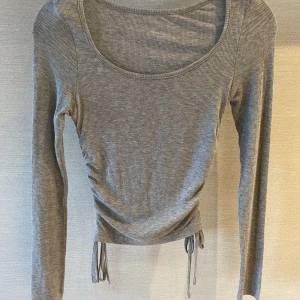 Tröja  - Långärmad stretchig grå tröja, drapperad i sidorna. Innearm längd på 47 cm och midjemått på 50 cm 