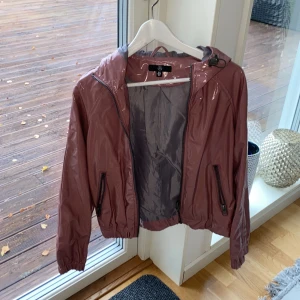 Regnjacka, glansig - Rosa/mauve glansig regnjacka från missguided. Relativt kort och har luva. Fint skick, endast använd någon enstaka gång.