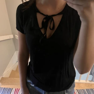 Svart T-shirt med knytning - V-ringad kortärmad blus med knytning man kan göra till rosett. Skönt material, viskos, som är perfekt för sommaren. Faller fint på kroppen. 